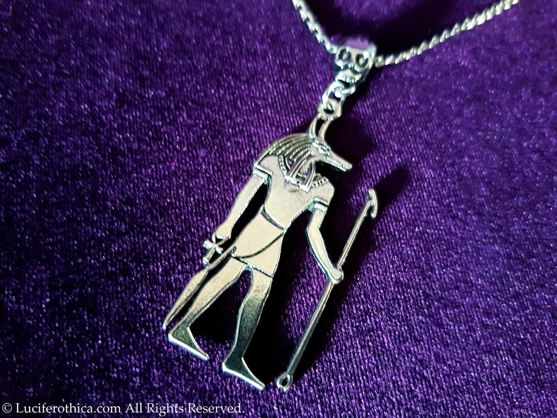 May include: Silver pendant necklace featuring an Egyptian god Anubis with a jackal head, holding a staff. The pendant is on a silver chain and is displayed on a purple velvet background.