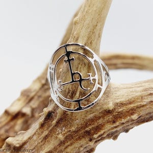 May include: Silver ring with a circular design featuring a stylized cross and other symbols. The ring is displayed on a piece of wood.