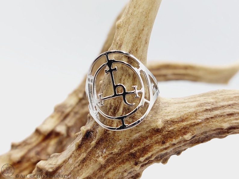 May include: Silver ring with a circular design featuring a stylized cross and other symbols. The ring is displayed on a piece of wood.