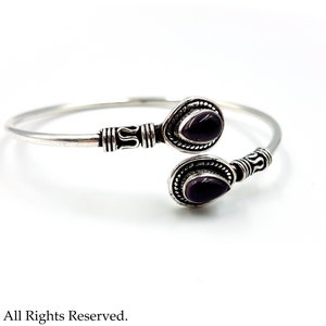 May include: A silver open cuff bracelet featuring two teardrop-shaped purple gemstones at the ends. The metal band displays intricate oxidized silver detailing and textured patterns near the settings of the two dark purple stones.