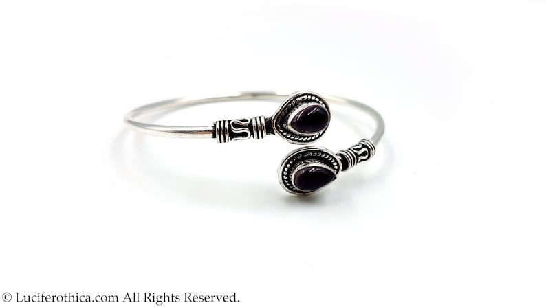 May include: A silver open cuff bracelet featuring two teardrop-shaped purple gemstones at the ends. The metal band displays intricate oxidized silver detailing and textured patterns near the settings of the two dark purple stones.