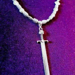 May include: A silver necklace with a sword pendant. The necklace is made of small, round beads and the sword is detailed with a pointed tip and a cross guard.