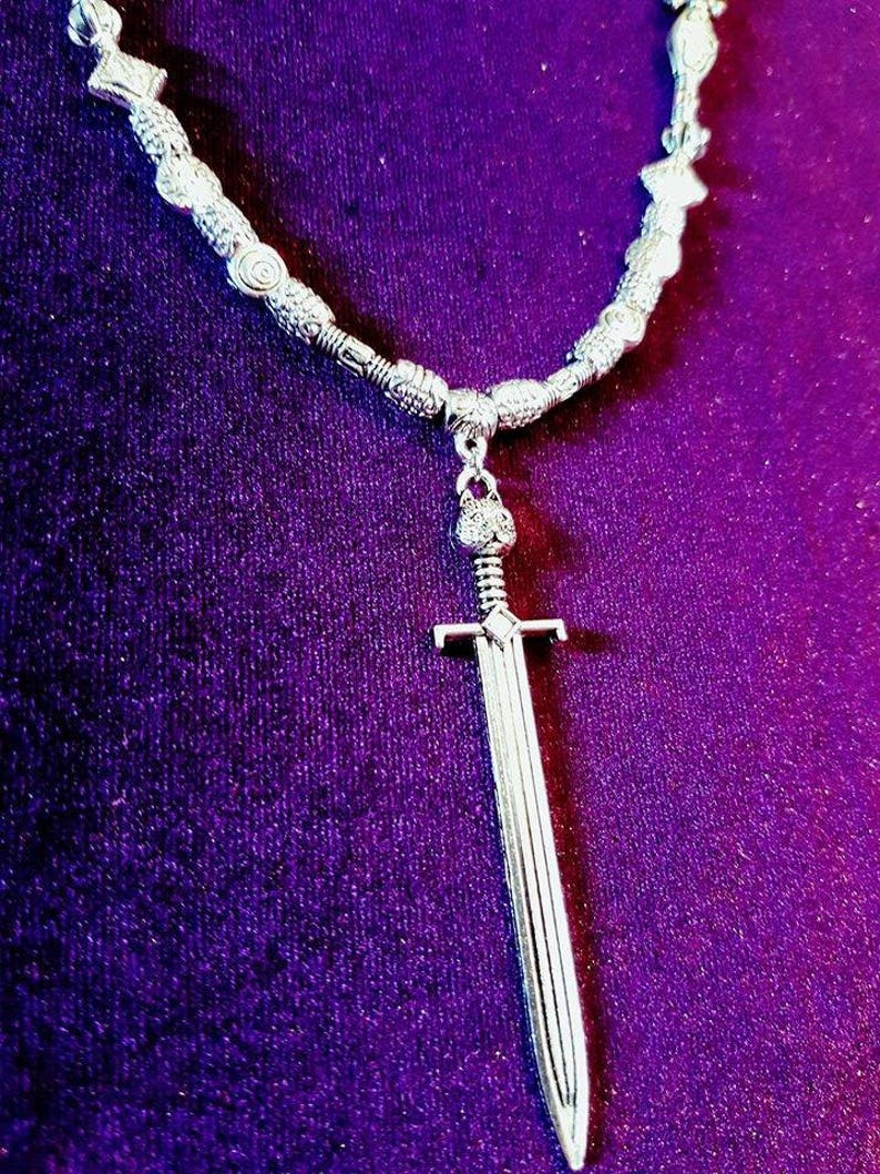 May include: A silver necklace with a sword pendant. The necklace is made of small, round beads and the sword is detailed with a pointed tip and a cross guard.