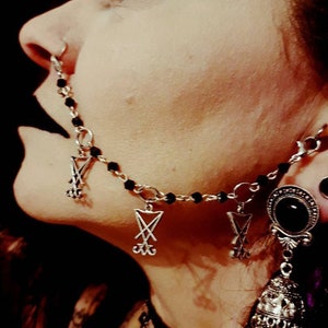 Könnte beinhalten: Eine silberne Nasenkette mit mehreren Pentagramm-Anhängern. Die Kette wird von einer Person mit dunklem Make-up und einem schwarzen Ohrring mit einem schwarzen Stein getragen.