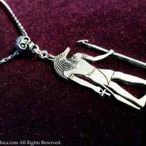 May include: Silver necklace with a pendant featuring an Anubis figure holding a staff. The pendant is attached to a silver chain with a skull bead.