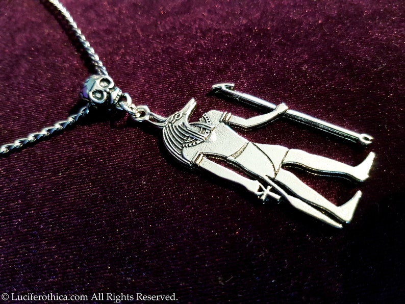 May include: Silver necklace with a pendant featuring an Anubis figure holding a staff. The pendant is attached to a silver chain with a skull bead.