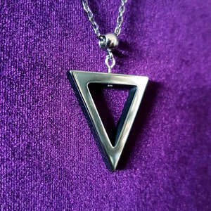 May include: A silver chain necklace with a triangle pendant. The triangle is hollow and has a silver border. The necklace is on a purple background.