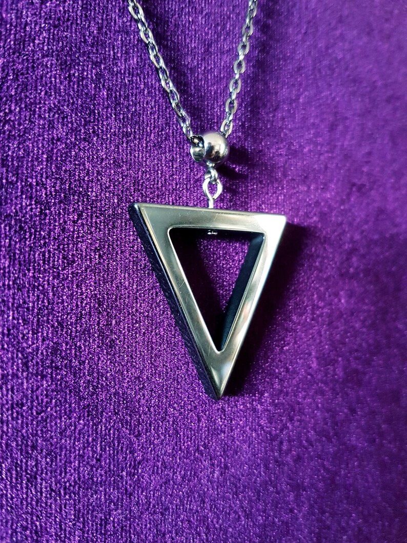 May include: A silver chain necklace with a triangle pendant. The triangle is hollow and has a silver border. The necklace is on a purple background.