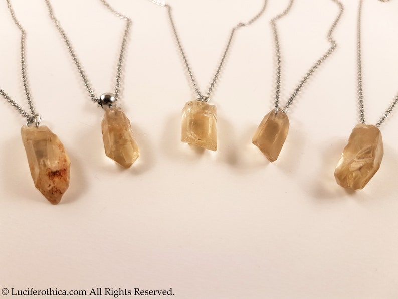 May include: Five silver necklaces with raw, light brown, crystal pendants. The necklaces are on a white background.