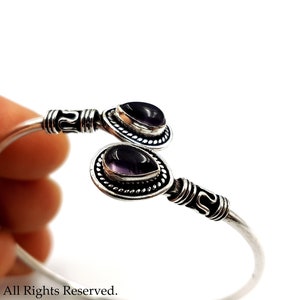 May include: A silver cuff bracelet featuring two teardrop-shaped purple amethyst stones set in ornate metal bezels. The band has a polished finish with decorative etched detailing near the ends, which hold the stones in an open-ended design.