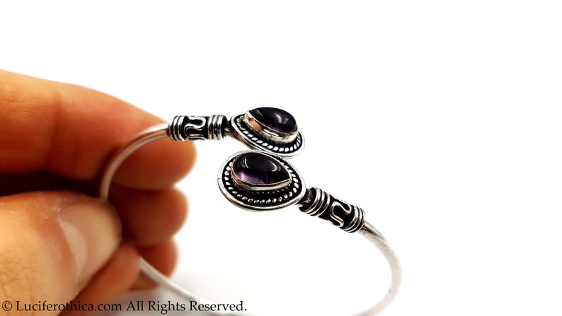 May include: A silver cuff bracelet featuring two teardrop-shaped purple amethyst stones set in ornate metal bezels. The band has a polished finish with decorative etched detailing near the ends, which hold the stones in an open-ended design.