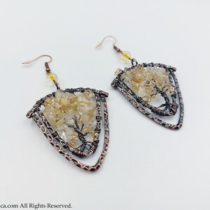May include: A pair of copper wire earrings featuring a tree design. The tree is crafted from small, clear and yellow citrine stones. The earrings are shaped like a triangle.