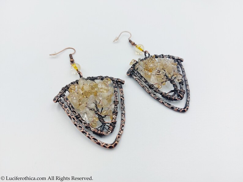 May include: A pair of copper wire earrings featuring a tree design. The tree is crafted from small, clear and yellow citrine stones. The earrings are shaped like a triangle.