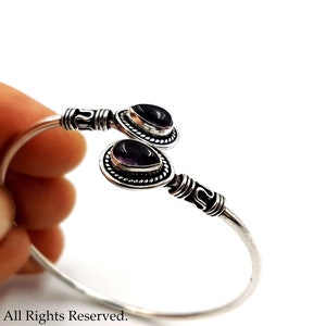 May include: A silver cuff bracelet featuring two teardrop-shaped purple amethyst gemstones set in decorative metal bezels. The band has a polished finish with intricate etched detailing near the stone settings at each open end of the bracelet.