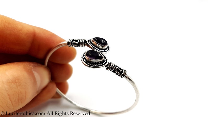 May include: A silver cuff bracelet featuring two teardrop-shaped purple amethyst gemstones set in decorative metal bezels. The band has a polished finish with intricate etched detailing near the stone settings at each open end of the bracelet.