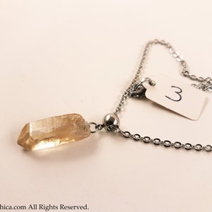 May include: A gold crystal pendant on a silver chain necklace. The pendant is a long, thin, pointed crystal. The chain has a small silver bead near the pendant.