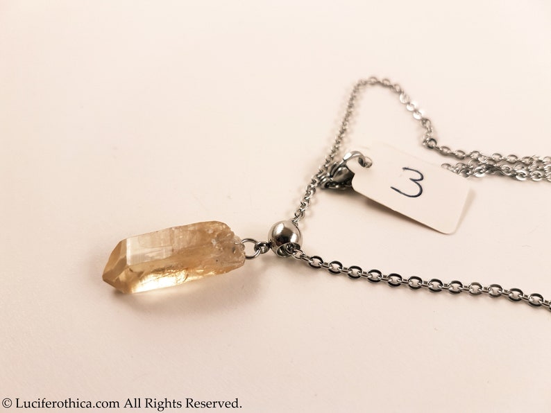 May include: A gold crystal pendant on a silver chain necklace. The pendant is a long, thin, pointed crystal. The chain has a small silver bead near the pendant.