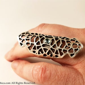 May include: A silver ring with a delicate, intricate design and a small blue gemstone. The ring is designed to be worn on the knuckle of the finger.