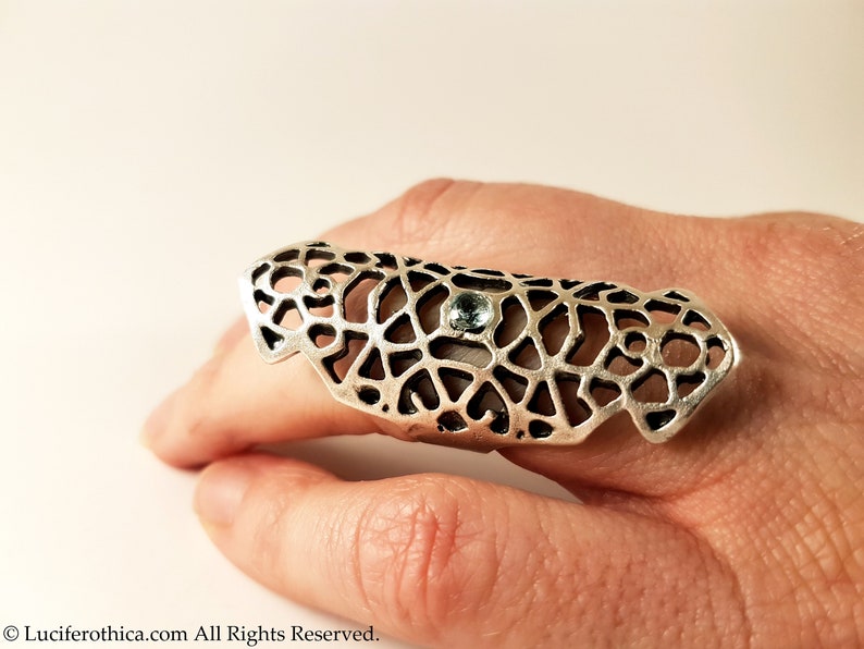 May include: A silver ring with a delicate, intricate design and a small blue gemstone. The ring is designed to be worn on the knuckle of the finger.