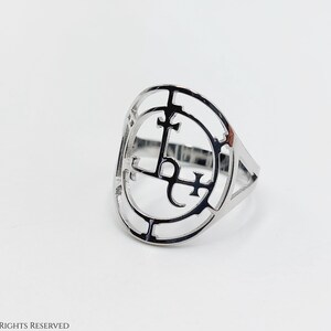 May include: A silver ring with a circular design featuring a stylized symbol with crosses and a letter L.