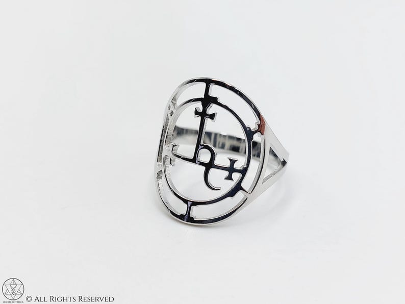 May include: A silver ring with a circular design featuring a stylized symbol with crosses and a letter L.