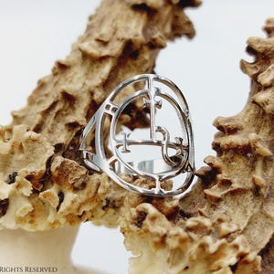 May include: A silver ring with a detailed design of a sigil. The ring is resting on a piece of natural, light brown, textured material.