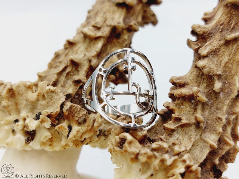 May include: A silver ring with a detailed design of a sigil. The ring is resting on a piece of natural, light brown, textured material.