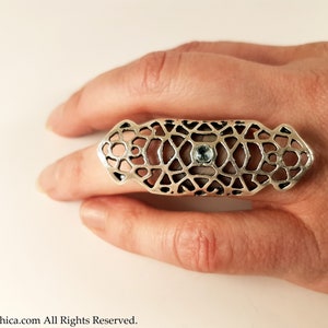 May include: A silver ring with a geometric design and a light blue gemstone in the center. The ring is worn on a finger.