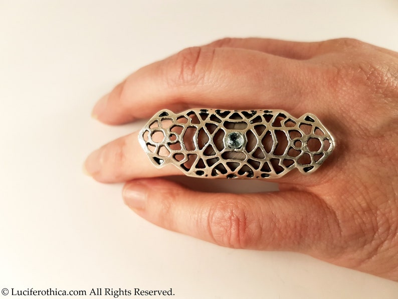 May include: A silver ring with a geometric design and a light blue gemstone in the center. The ring is worn on a finger.