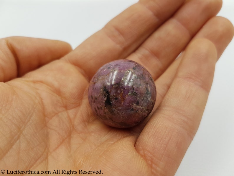May include: A pink and purple sphere made of polished ruby in a hand. The sphere has a rough, textured surface.