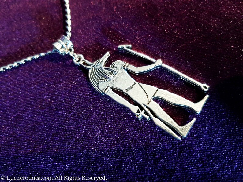 May include: Silver necklace featuring a pendant depicting an Egyptian god with a jackal head, holding a staff. The pendant is detailed and intricate.