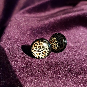 Puede incluir: Un par de pendientes redondos con borde negro y diseño de estampado de leopardo. Los pendientes están sobre una superficie morada brillante.