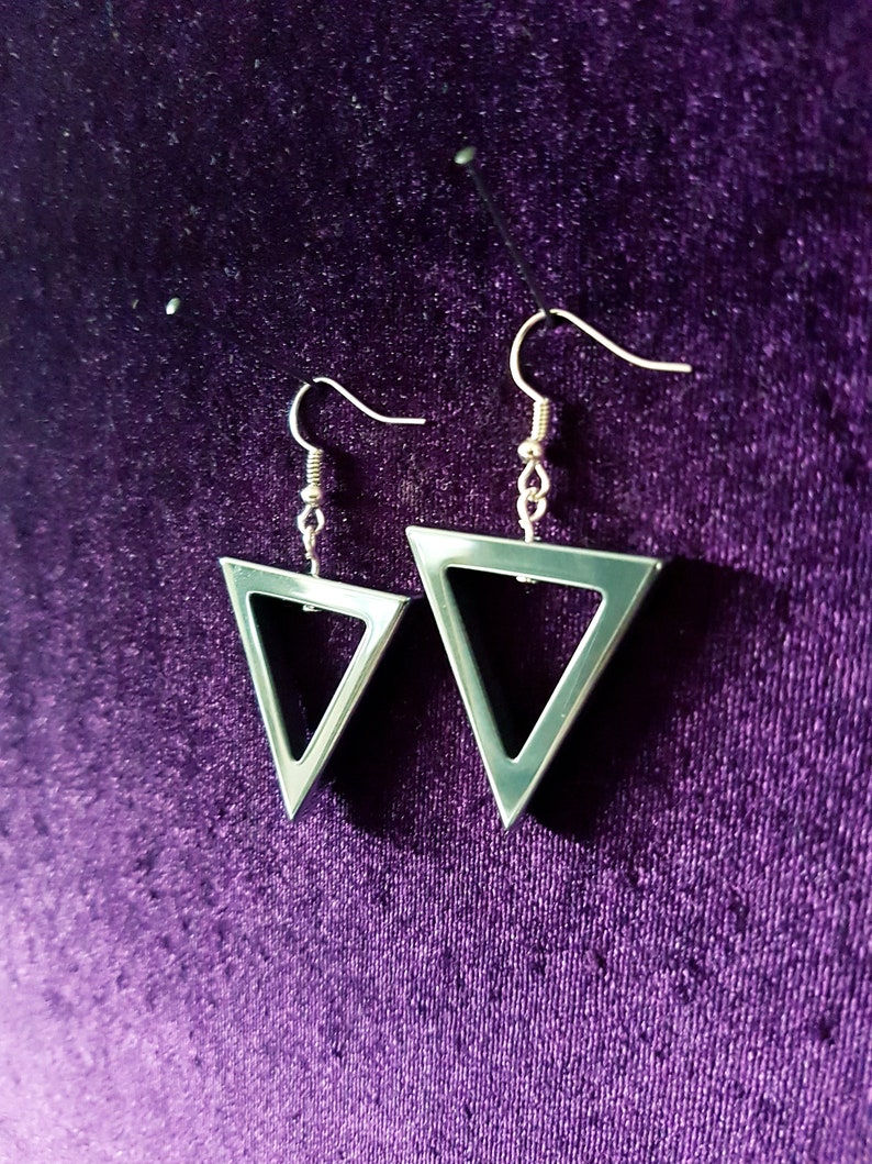 May include: A pair of silver triangle earrings with a hollow center. The earrings are hanging from a purple velvet background.