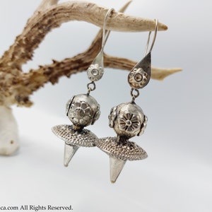 May include: A pair of silver dangle earrings with a round, floral-embossed bead and a pointed, textured disc.