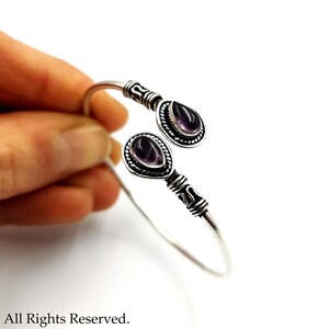 May include: A silver open cuff bracelet featuring two teardrop-shaped purple amethyst stones set in decorative metal bezels. The band has a twisted wire texture near the stone settings and a smooth, polished finish along the rest of the metal.