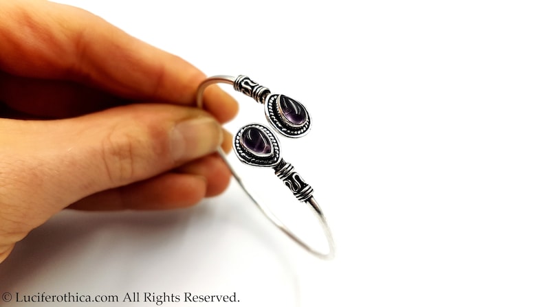 May include: A silver open cuff bracelet featuring two teardrop-shaped purple amethyst stones set in decorative metal bezels. The band has a twisted wire texture near the stone settings and a smooth, polished finish along the rest of the metal.