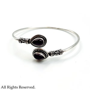 May include: A silver open cuff bracelet featuring two teardrop-shaped dark purple stones at the ends. The metal band has decorative etched detailing near the settings, which are framed by a twisted rope pattern. The text Luciferthica.com All Rights Reserved is visible at the bottom.
