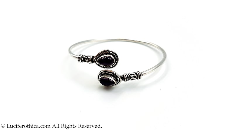 May include: A silver open cuff bracelet featuring two teardrop-shaped dark purple stones at the ends. The metal band has decorative etched detailing near the settings, which are framed by a twisted rope pattern. The text Luciferthica.com All Rights Reserved is visible at the bottom.