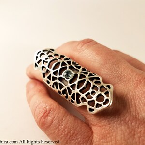 May include: A silver ring with a delicate, intricate design and a light blue gemstone in the center. The ring is worn on a finger and is a statement piece.