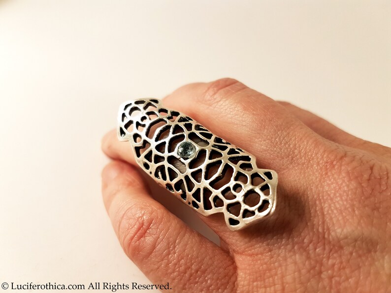 May include: A silver ring with a delicate, intricate design and a light blue gemstone in the center. The ring is worn on a finger and is a statement piece.