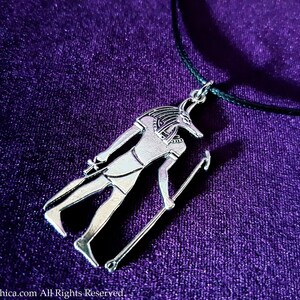May include: Silver pendant necklace featuring an Anubis figure with a jackal head, standing with a staff, on a black cord against a purple background.