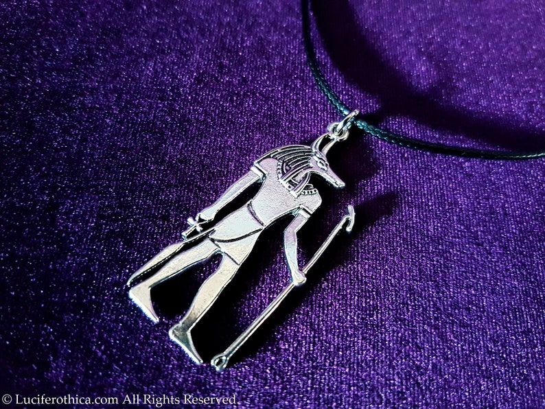 May include: Silver pendant necklace featuring an Anubis figure with a jackal head, standing with a staff, on a black cord against a purple background.