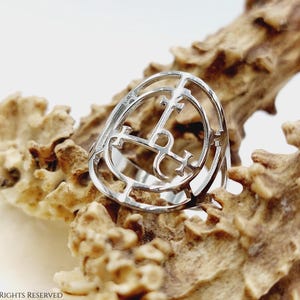 May include: A silver ring with a circular design featuring a cross and other symbols. The ring is displayed on a textured, light brown surface.