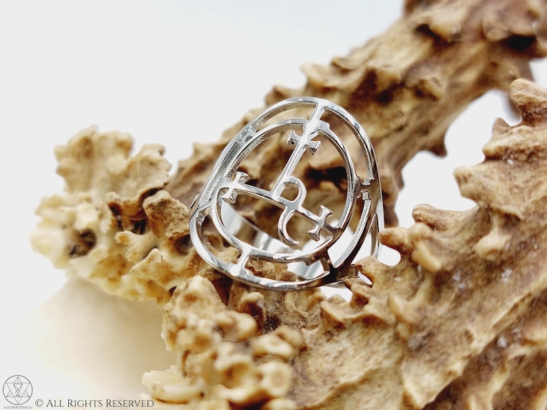 May include: A silver ring with a circular design featuring a cross and other symbols. The ring is displayed on a textured, light brown surface.