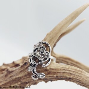 K&ouml;nnte beinhalten: Ein silberner Ring mit einem detaillierten Rosenmotiv. Die Rose befindet sich auf einem Ranken mit Bl&auml;ttern. Der Ring wird auf einem St&uuml;ck Geweih gezeigt.
