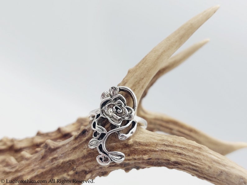 K&ouml;nnte beinhalten: Ein silberner Ring mit einem detaillierten Rosenmotiv. Die Rose befindet sich auf einem Ranken mit Bl&auml;ttern. Der Ring wird auf einem St&uuml;ck Geweih gezeigt.