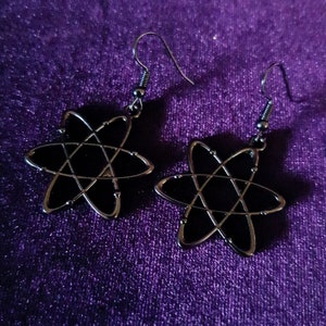 Może przedstawiać: Para czarnych i srebrnych kolczyków w kształcie atomu. Kolczyki mają ciemne, błyszczące centrum ze srebrnym obrysem. Haczyki są srebrne. Kolczyki są prezentowane na fioletowej aksamitnej powierzchni.