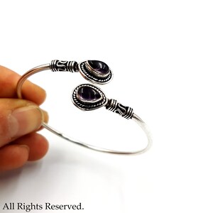 May include: A silver cuff bracelet featuring two teardrop-shaped purple amethyst stones set at each end. The metal band displays intricate oxidized detailing near the stones, which are framed by a beaded border. The design is open-ended.