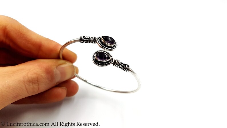 May include: A silver cuff bracelet featuring two teardrop-shaped purple amethyst stones set at each end. The metal band displays intricate oxidized detailing near the stones, which are framed by a beaded border. The design is open-ended.