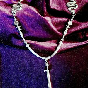 May include: A silver necklace with a sword pendant and two snake charms. The necklace is on a purple velvet background.